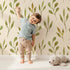 Lovely child standing in front of Whimsy Tots Whimsy Leaf Branch Wallpaper in Cream with leaf, stem, greenery, branch in design.