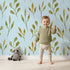 Lovely child standing in front of Whimsy Tots Whimsy Leaf Branch Wallpaper in Sky with leaf, stem, greenery, branch in design.