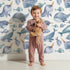 Lovely child standing in front of Whimsy Tots Sleepy Ocean Friends Wallpaper in White with whale, narwhal, fish, sea in design.