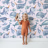 Lovely child standing in front of Whimsy Tots Sleepy Ocean Friends Wallpaper in Blush Pink with whale, narwhal, fish, sea in design.