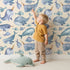 Lovely child standing in front of Whimsy Tots Sleepy Ocean Friends Wallpaper in Cream with whale, narwhal, fish, sea in design.