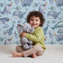 Lovely child standing in front of Whimsy Tots Sleepy Ocean Friends Wallpaper in Sky with whale, narwhal, fish, sea in design.