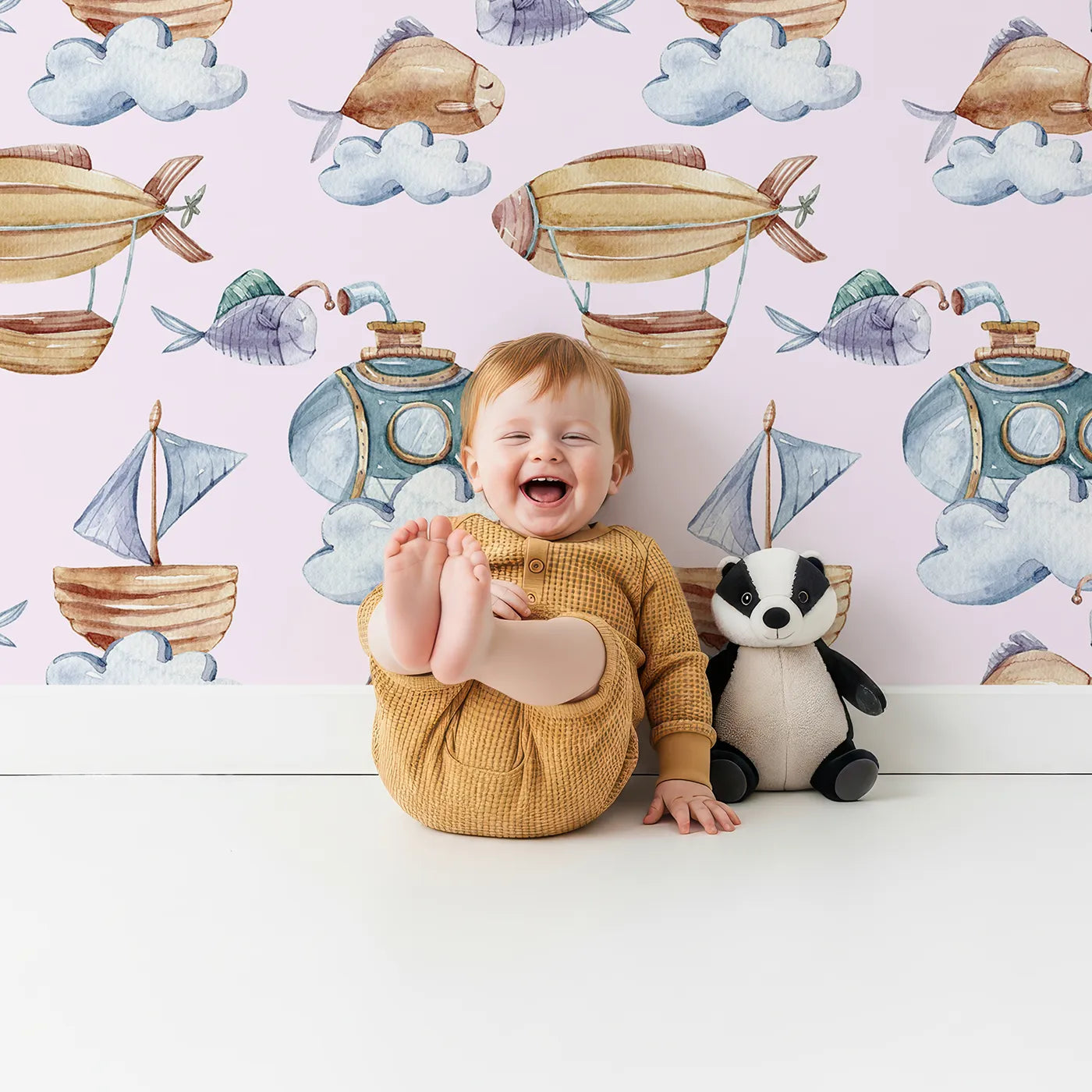 Lovely child standing in front of Whimsy Tots Sky Explorers Wallpaper in Blush Pink with fish, submarine, balloon, ship, sky, cloud in design.