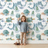Lovely child standing in front of Whimsy Tots Ocean Daydream Creatures Wallpaper in White with whale, fish, turtle, octopus, dinosaur, seaweed in design.