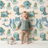 Lovely child standing in front of Whimsy Tots Ocean Daydream Creatures Wallpaper in Cream with whale, fish, turtle, octopus, dinosaur, seaweed in design.