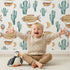 Lovely child standing in front of Whimsy Tots Desert Balloon Voyage Wallpaper in White with airship, cactus, moon, balloon in design.