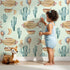 Lovely child standing in front of Whimsy Tots Desert Balloon Voyage Wallpaper in Mint Green with airship, cactus, moon, balloon in design.