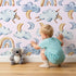 Lovely child standing in front of Whimsy Tots Sleepy Skies Wallpaper in Blush Pink with cloud, moon, rainbow, sheep in design.