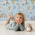Lovely child standing in front of Whimsy Tots Sleepy Skies Wallpaper in Blue with cloud, moon, rainbow, sheep in design.