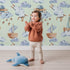 Lovely child standing in front of Whimsy Tots Cloud Sailor Dreams Wallpaper in Mint Green with ship, cloud, sheep, star, sail in design.