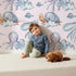 Lovely child standing in front of Whimsy Tots Ocean Dreams Wallpaper in Blush Pink with cloud, turtle, octopus, squid, fish in design.