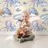 Lovely child standing in front of Whimsy Tots Ocean Dreams Wallpaper in Cream with cloud, turtle, octopus, squid, fish in design.