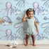 Lovely child standing in front of Whimsy Tots Ocean Dreams Wallpaper in Blue with cloud, turtle, octopus, squid, fish in design.