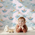 Lovely child standing in front of Whimsy Tots Whale Boat Parade Wallpaper in Pink with whale, submarine, boat, octopus, anchor, flag in design.