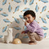 Lovely child standing in front of Whimsy Tots Little Swimmer Fish Wallpaper in White with fish in design.