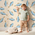 Lovely child standing in front of Whimsy Tots Little Swimmer Fish Wallpaper in Cream with fish in design.
