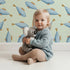 Lovely child standing in front of Whimsy Tots Little Swimmer Fish Wallpaper in Mint Green with fish, little, swimmer, wallpaper in design.