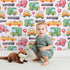 Lovely child standing in front of Whimsy Tots Little Car Parade Wallpaper in Pink with car, sign, road in design.