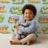 Lovely child standing in front of Whimsy Tots Cottage Garden Village Wallpaper in Blue with tree, bush, house in design.