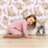 Lovely child standing in front of Whimsy Tots Dried Bloom Cascade Wallpaper in Pink with flower, palm, leaf in design.