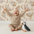 Lovely child standing in front of Whimsy Tots Dried Bloom Cascade Wallpaper in Cream with flower, palm, leaf in design.