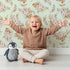 Lovely child standing in front of Whimsy Tots Dried Bloom Cascade Wallpaper in Mint Green with flower, palm, leaf in design.