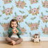 Lovely child standing in front of Whimsy Tots Rustic Blush Bouquet Wallpaper in Blue with flower, palm, leaf in design.