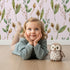 Lovely child standing in front of Whimsy Tots Woodland Forage Sprigs Wallpaper in Pink with leaf, pod, sprig in design.