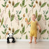 Lovely child standing in front of Whimsy Tots Woodland Forage Sprigs Wallpaper in Cream with leaf, pod, sprig in design.
