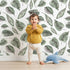 Lovely child standing in front of Whimsy Tots Monsoon Leaf Drift Wallpaper in White with leaf in design.