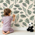 Lovely child standing in front of Whimsy Tots Monsoon Leaf Drift Wallpaper in Cream with leaf in design.