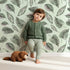 Lovely child standing in front of Whimsy Tots Monsoon Leaf Drift Wallpaper in Mint Green with monsoon, leaf, drift, wallpaper in design.