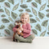 Lovely child standing in front of Whimsy Tots Monsoon Leaf Drift Wallpaper in Blue with leaf in design.