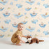 Lovely child standing in front of Whimsy Tots Whale Submarine Adventure Wallpaper in Beige with whale, submarine, fish, anchor, octopus, bunting in design.