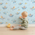 Lovely child standing in front of Whimsy Tots Whale Submarine Adventure Wallpaper in Sky Blue with whale, submarine, fish, anchor, octopus, bunting in design.