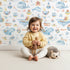 Lovely child standing in front of Whimsy Tots Whale Mail Wallpaper in White with whale, fish, octopus, boat, lighthouse, anchor, bird, mug, watermelon, letter in design.