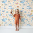 Lovely child standing in front of Whimsy Tots Whale Mail Wallpaper in Peach with whale, fish, octopus, boat, lighthouse, anchor, bird, mug, watermelon, letter in design.