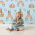 Lovely child standing in front of Whimsy Tots Rainbow Castle Dreams Wallpaper in Blue with castle, rainbow, moon, cloud, star, net in design.
