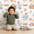 Lovely child standing in front of Whimsy Tots Dreamland Friends Wallpaper in White with bird, bunny, cloud, balloon, star in design.