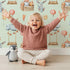 Lovely child standing in front of Whimsy Tots Dreamland Friends Wallpaper in Mint Green with bird, bunny, cloud, balloon, star in design.