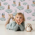Lovely child standing in front of Whimsy Tots Castle Cloud Drift Wallpaper in Pink with castle, cloud, moon in design.