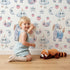 Lovely child standing in front of Whimsy Tots Starlight Narwhal Parade Wallpaper in White with narwhal, unicorn, moon, star, cloud, heart, fruit in design.