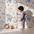 Lovely child standing in front of Whimsy Tots Starlight Narwhal Parade Wallpaper in Beige with narwhal, unicorn, moon, star, cloud, heart, fruit in design.