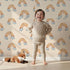 Lovely child standing in front of Whimsy Tots Sunset Rainbows Wallpaper in Beige with rainbow, cloud in design.