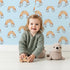 Lovely child standing in front of Whimsy Tots Sunset Rainbows Wallpaper in Blue with rainbow, cloud in design.