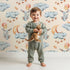 Lovely child standing in front of Whimsy Tots Moonlight Bunny Dreams Wallpaper in Beige with rabbit, balloon, moon, star, cloud in design.