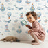Lovely child standing in front of Whimsy Tots Maritime Whimsy Wallpaper in White with whale, octopus, boat, sailor, cloud, bulb, bottle in design.