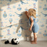 Lovely child standing in front of Whimsy Tots Maritime Whimsy Wallpaper in Cream with whale, octopus, boat, sailor, cloud, bulb, bottle in design.