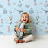 Lovely child standing in front of Whimsy Tots Maritime Whimsy Wallpaper in Blue with whale, octopus, boat, sailor, cloud, bulb, bottle in design.