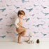 Lovely child standing in front of Whimsy Tots Whale Cloud Voyage Wallpaper in Blush Pink with whale, cloud, splash in design.